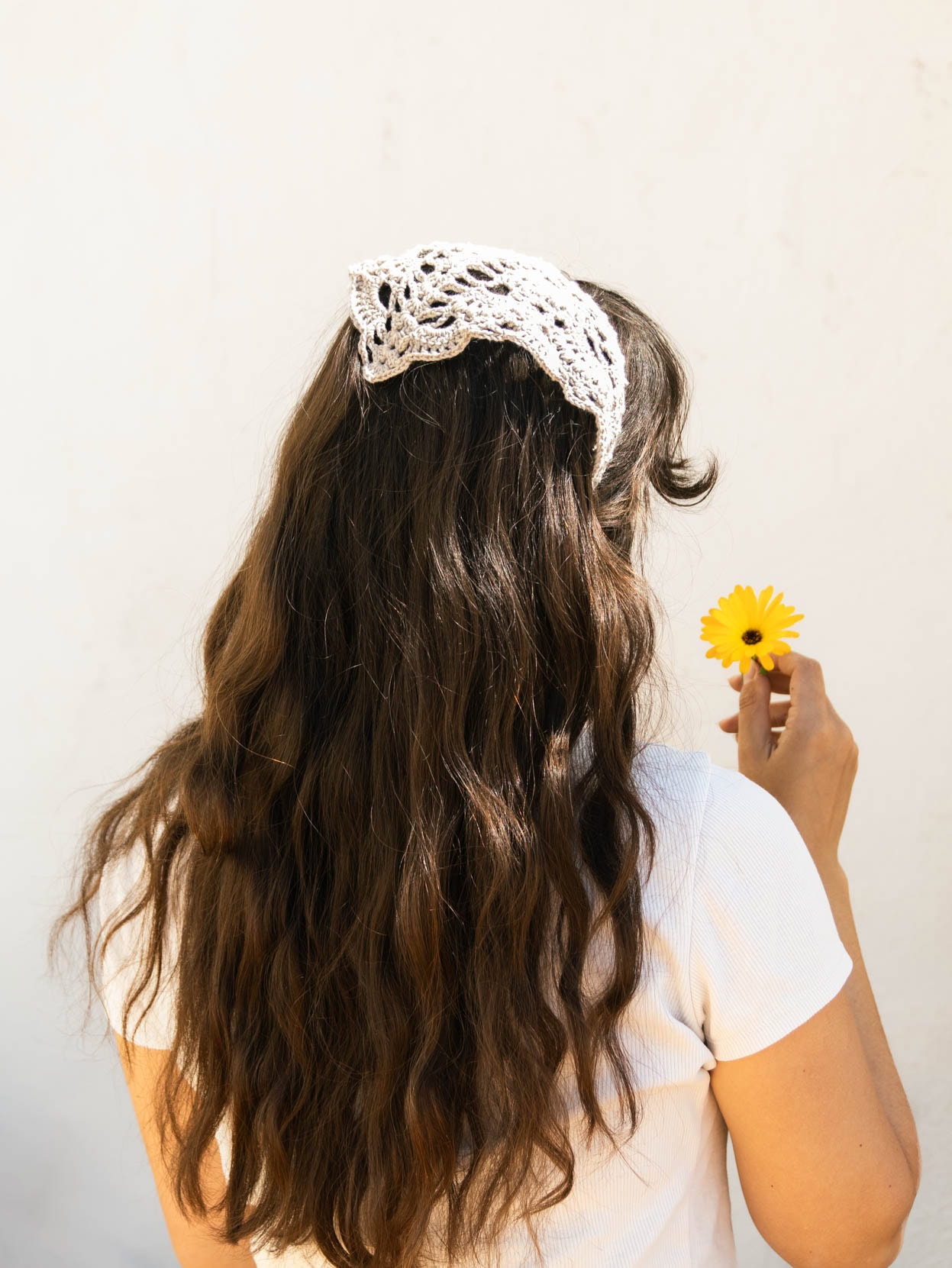 Back view of a cream crochet bandana worn over long wavy hair while holding a yellow flower